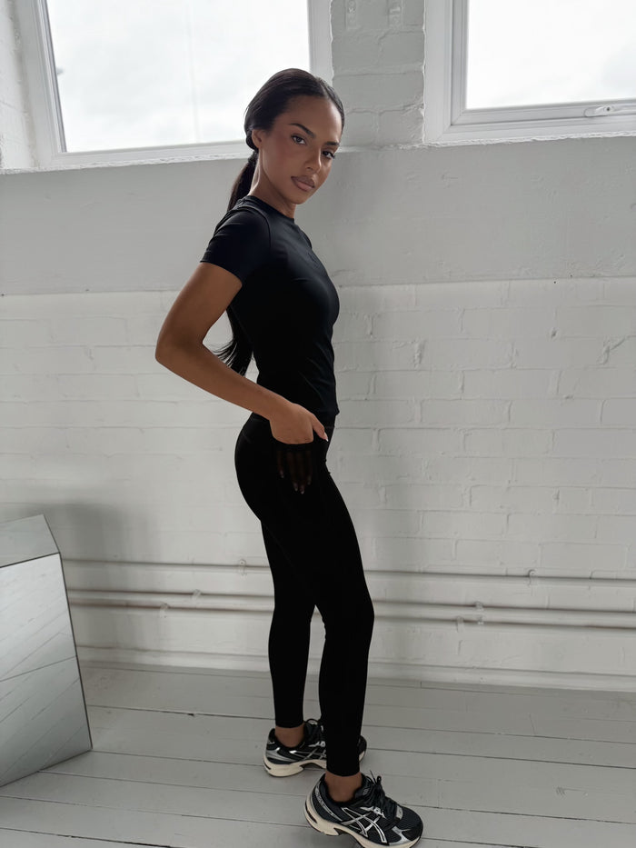 Black Define Sculpt Tee worn with black leggings, modelled from the side against a white brick wall