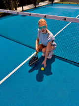 A young girl wearing oversized reflective sunglasses crouches on a bright blue tennis court near the net, holding a paddle racket and reaching for a yellow tennis ball.
